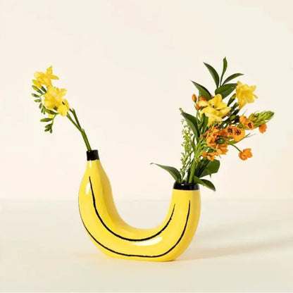 Yellow resin banana bunch vase holding dried flower stems on wooden shelf
