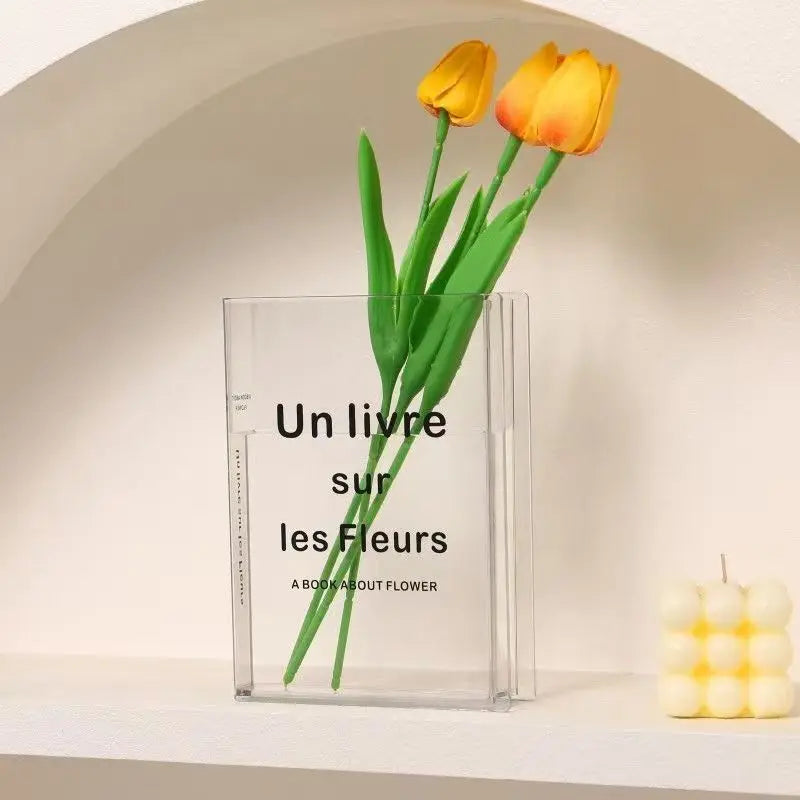 Transparent acrylic open book vase holding single white flower bloom on desk surface