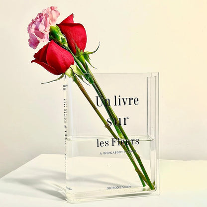 Clear acrylic book-shaped vase with fresh flowers displayed between books on bookshelf